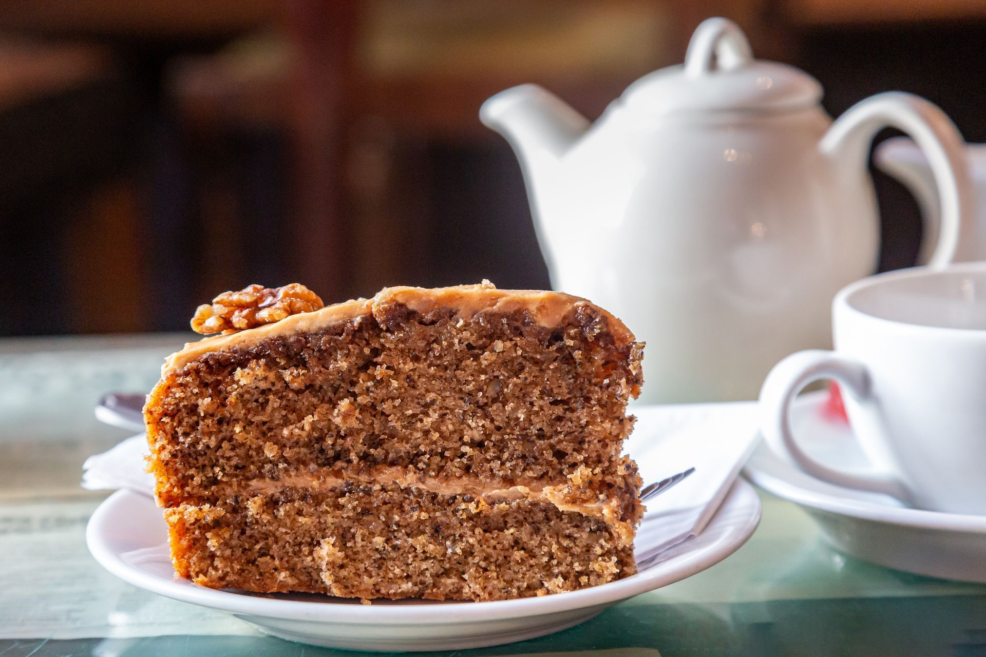 A slice of cake is on a plate next to a cup of coffee and a teapot.