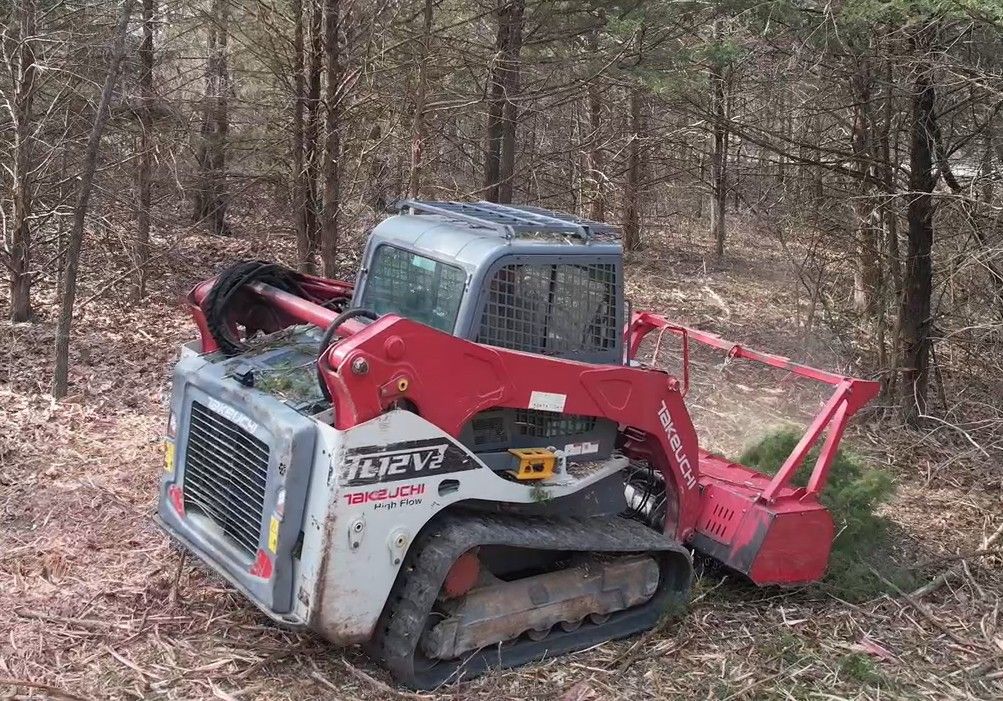 Skid steer with raised bucket of logs clearing debris in a muddy field, surrounded by trees under a cloudy sky.