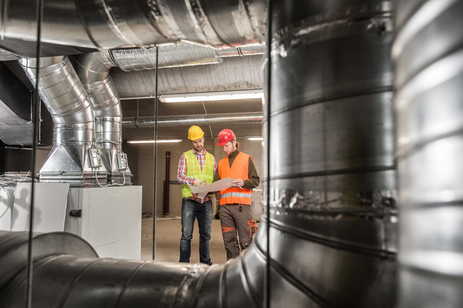 Two construction workers are looking at a blueprint in a room with pipes.