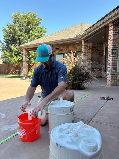 Man in a hat pouring from a bottle into a red bucket on a patio, next to two barrels and a green hose.