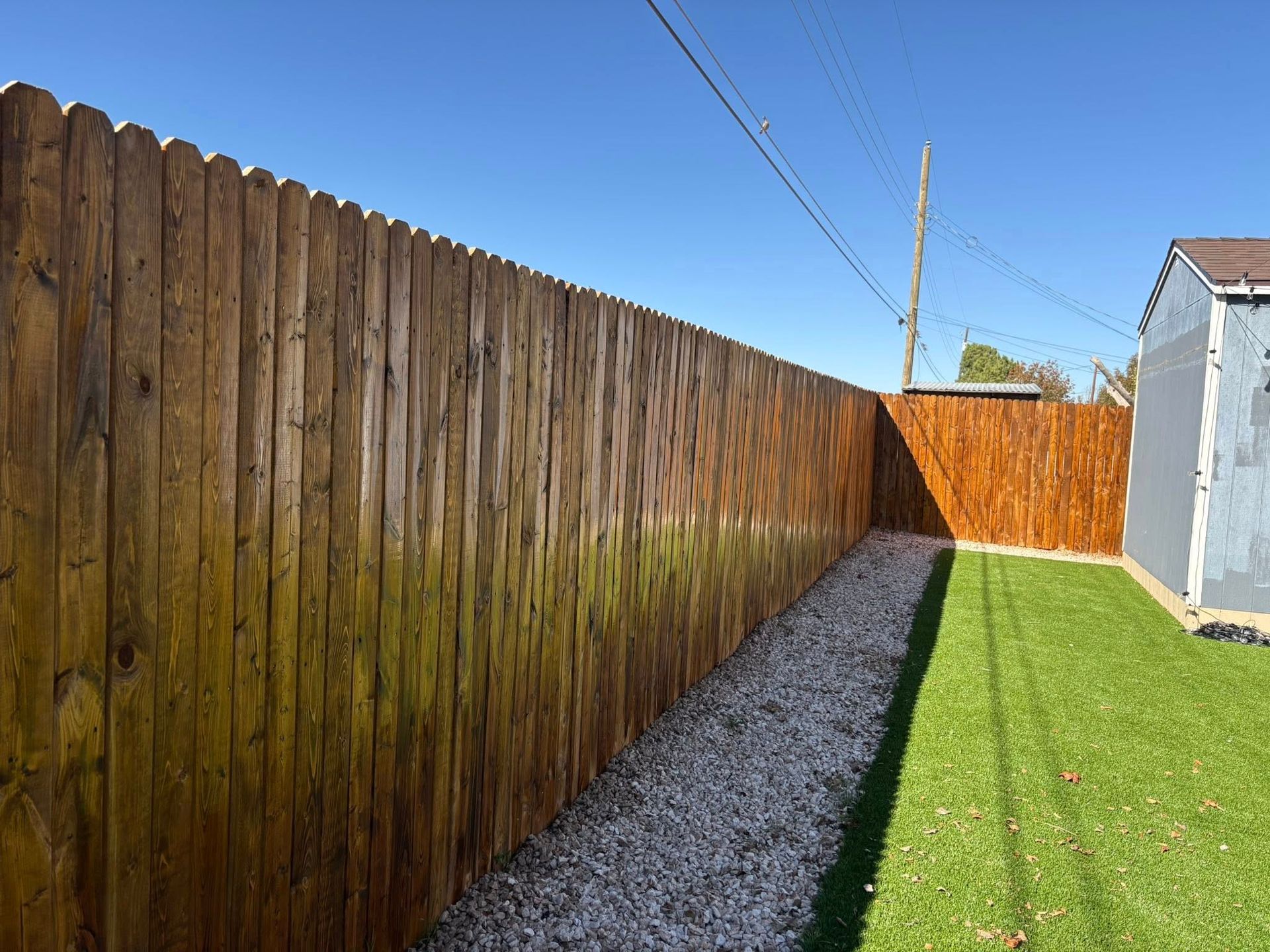 Wooden fence stained brown, gravel path, and green lawn against a blue sky.