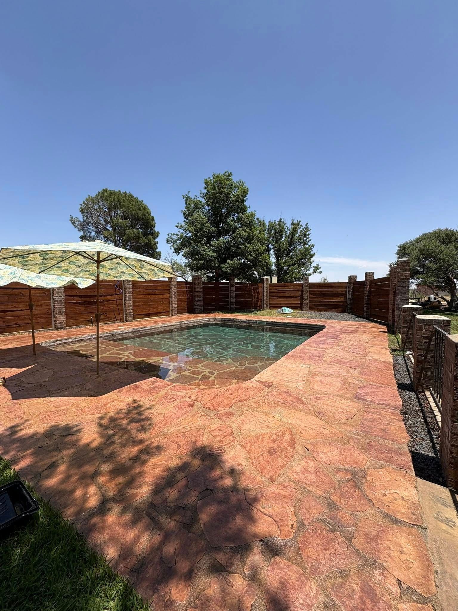 Pool surrounded by reddish-brown paving, with a fence and trees against a blue sky; an umbrella stands next to the water.