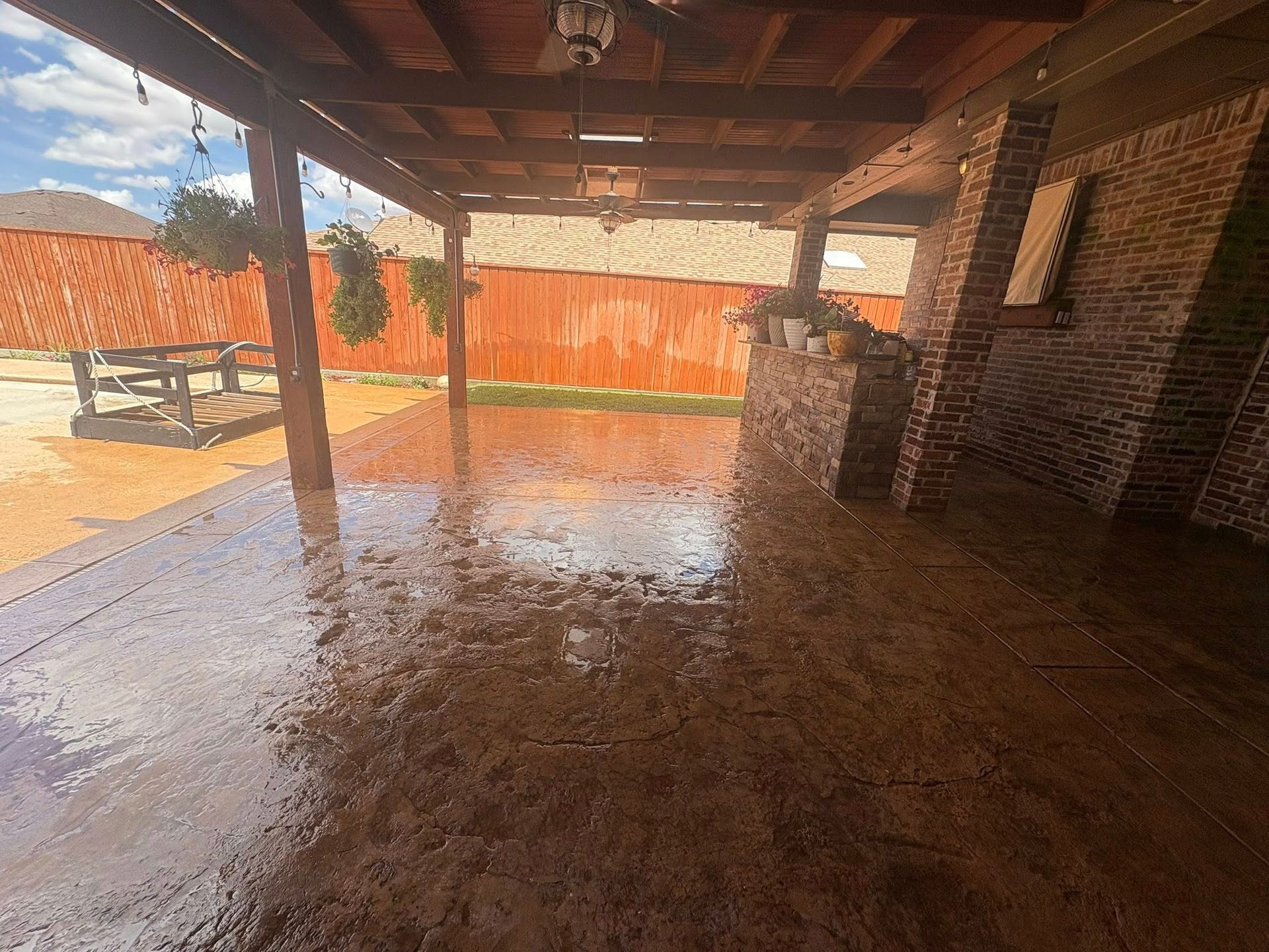Covered patio with wet, brown stamped concrete, brick accents, and wooden beams.