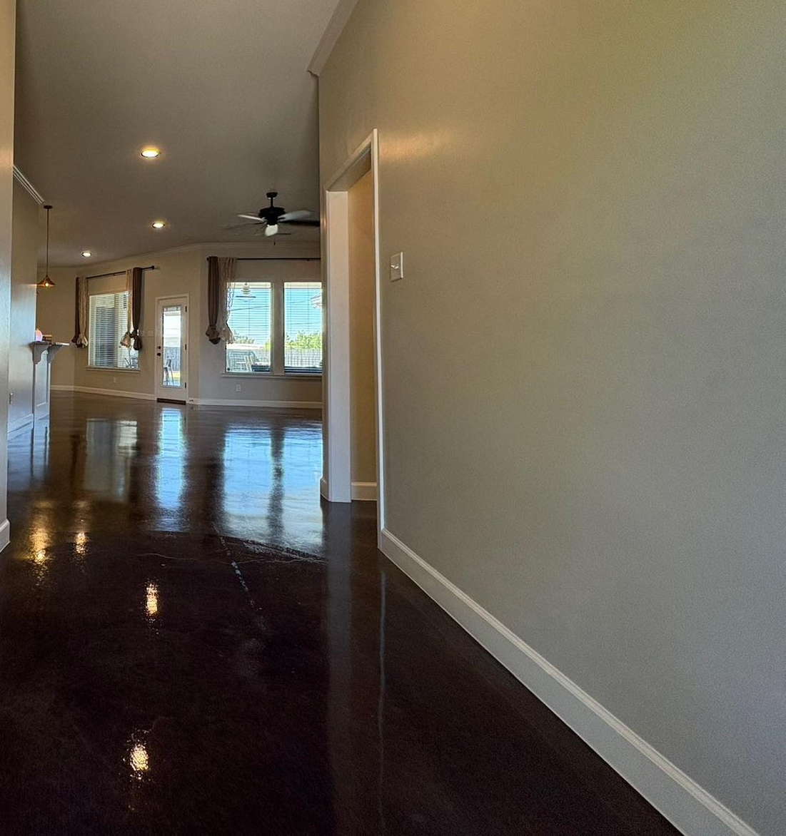 A long hallway with glossy brown floor leads to a bright living area. Beige walls, white trim, and recessed lighting.