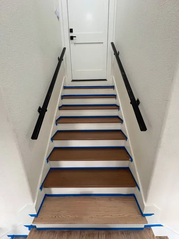 Staircase with wooden steps and black handrails, painted white walls, blue painter's tape along edges, leading to a white door.