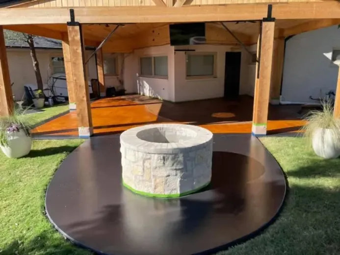Fire pit on a black circular platform, in a yard with grass, orange concrete patio, and covered wooden structure.