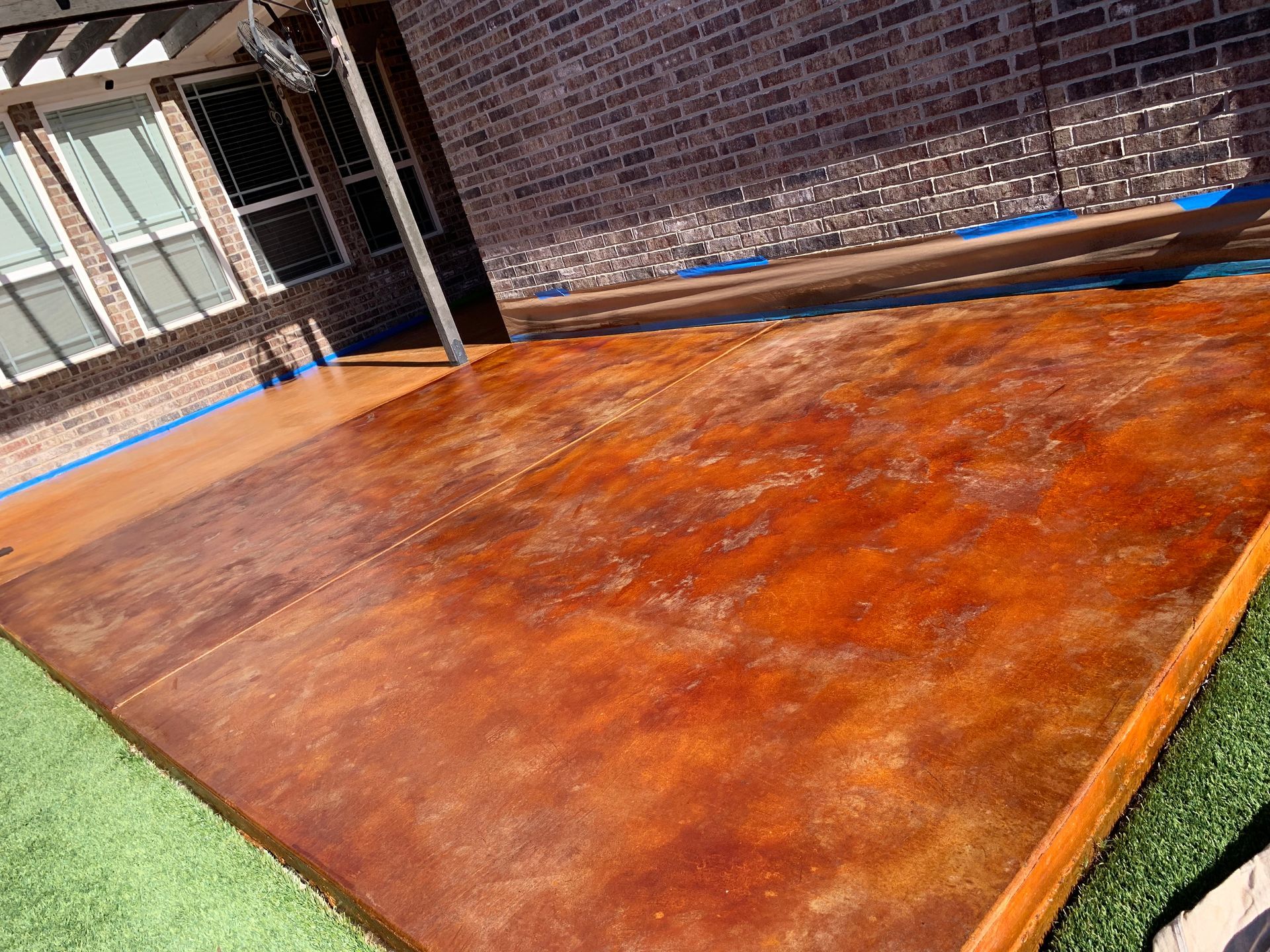 Brown stained concrete patio next to a brick wall and green turf, with blue painter's tape visible.