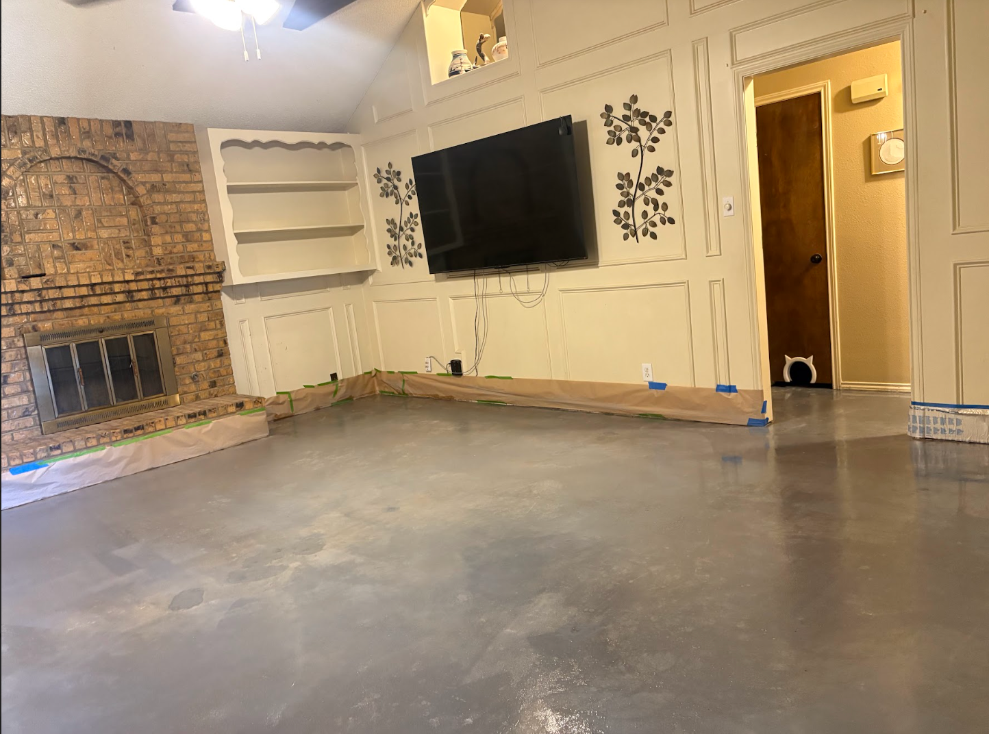 A living room with a brick fireplace, built-in shelving, wall-mounted television, and freshly painted gray concrete floors.