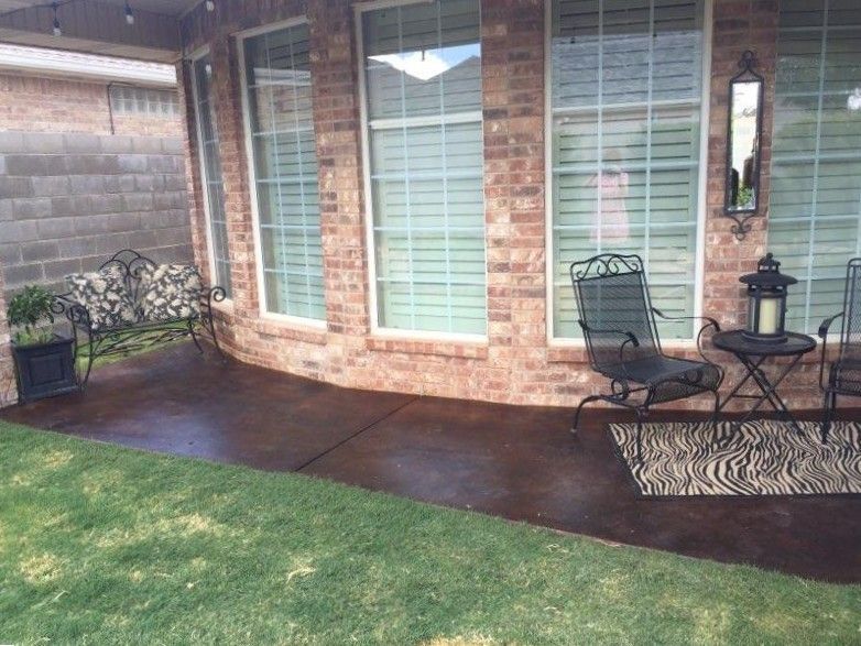 Brown concrete patio with outdoor furniture, next to a brick house and green lawn.