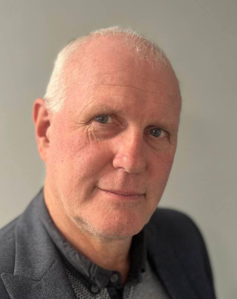 Man in a blazer, looking at the camera with a slight smile, against a light gray background.