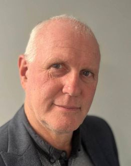 Man in a blazer, looking at the camera with a slight smile, against a light gray background.
