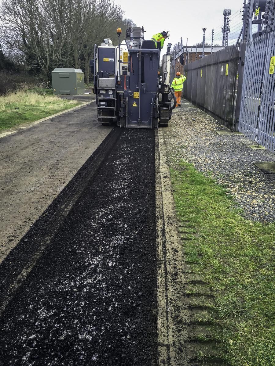 Road paving machine laying fresh asphalt on a narrow road, with workers in safety vests nearby.