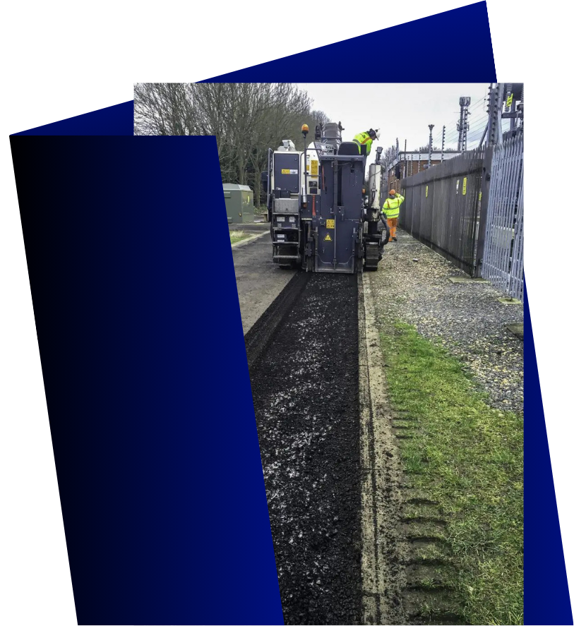 Asphalt paving machine laying blacktop in a narrow trench beside a fence. Worker in high-vis vest.