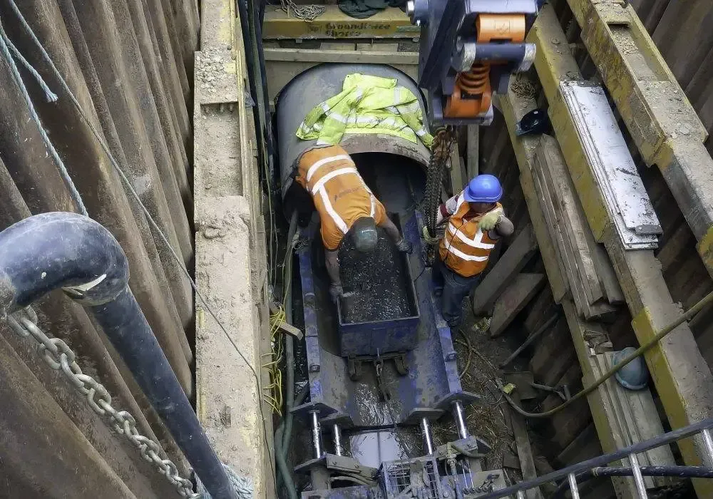 Workers inside a trench, installing a pipe. One wears a helmet and orange vest.