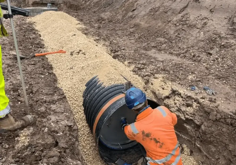 Construction worker in orange coveralls installing drainage pipe in trench lined with gravel.
