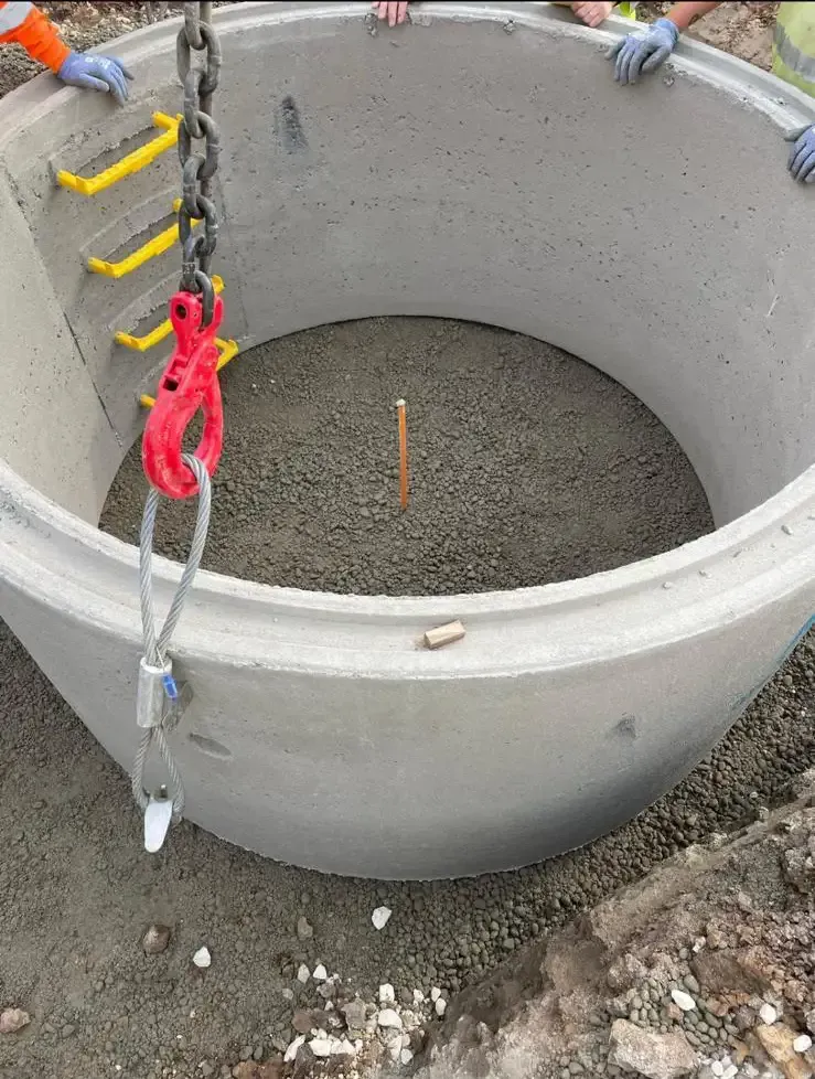 Concrete manhole being lowered into position with a chain hoist, visible safety rungs inside.