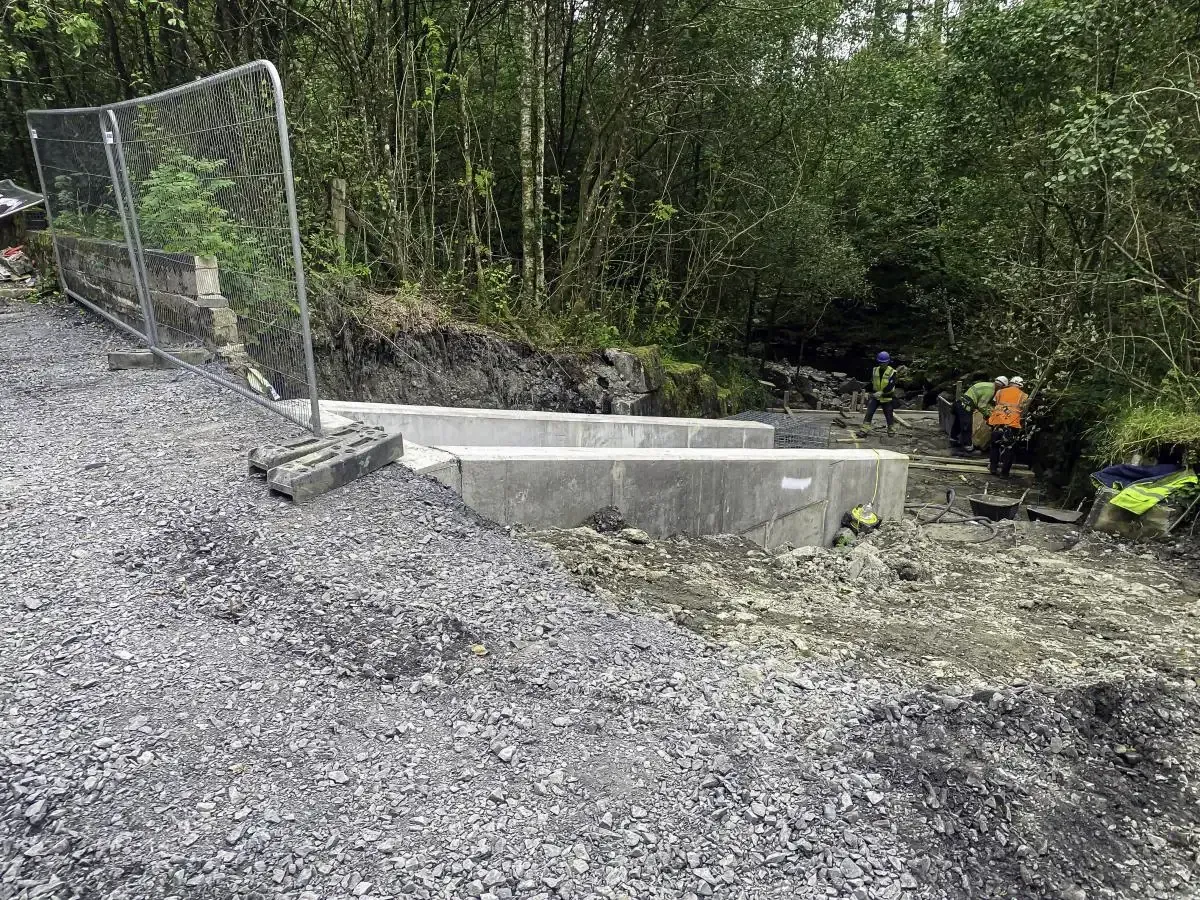 Concrete construction site in a wooded area; workers visible. Grey gravel ground, grey fence, and concrete.