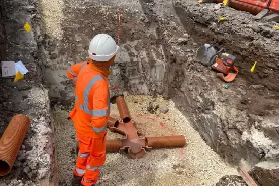 Construction worker in orange safety gear inspects brown pipes in a trench. Saw and flags are visible.