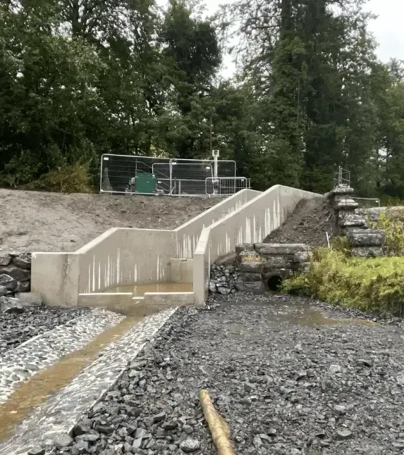Concrete fish ladder construction in a gravel-lined stream, next to forested area and structure.
