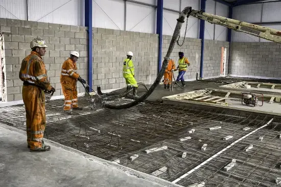 Construction workers pouring concrete on a wire mesh foundation inside a large building, using a pump.