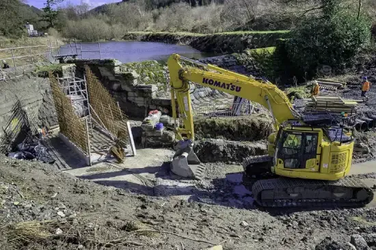 Yellow Komatsu excavator at a construction site near water, with rebar and a partially built structure.