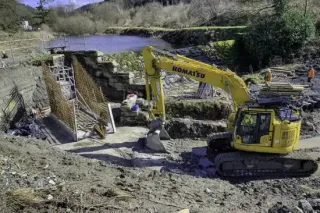Yellow Komatsu excavator at a construction site near water, with rebar and a partially built structure.