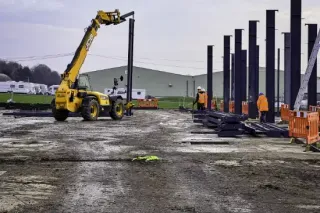 Construction site with a yellow telescopic handler lifting a black steel beam; workers in orange vests.