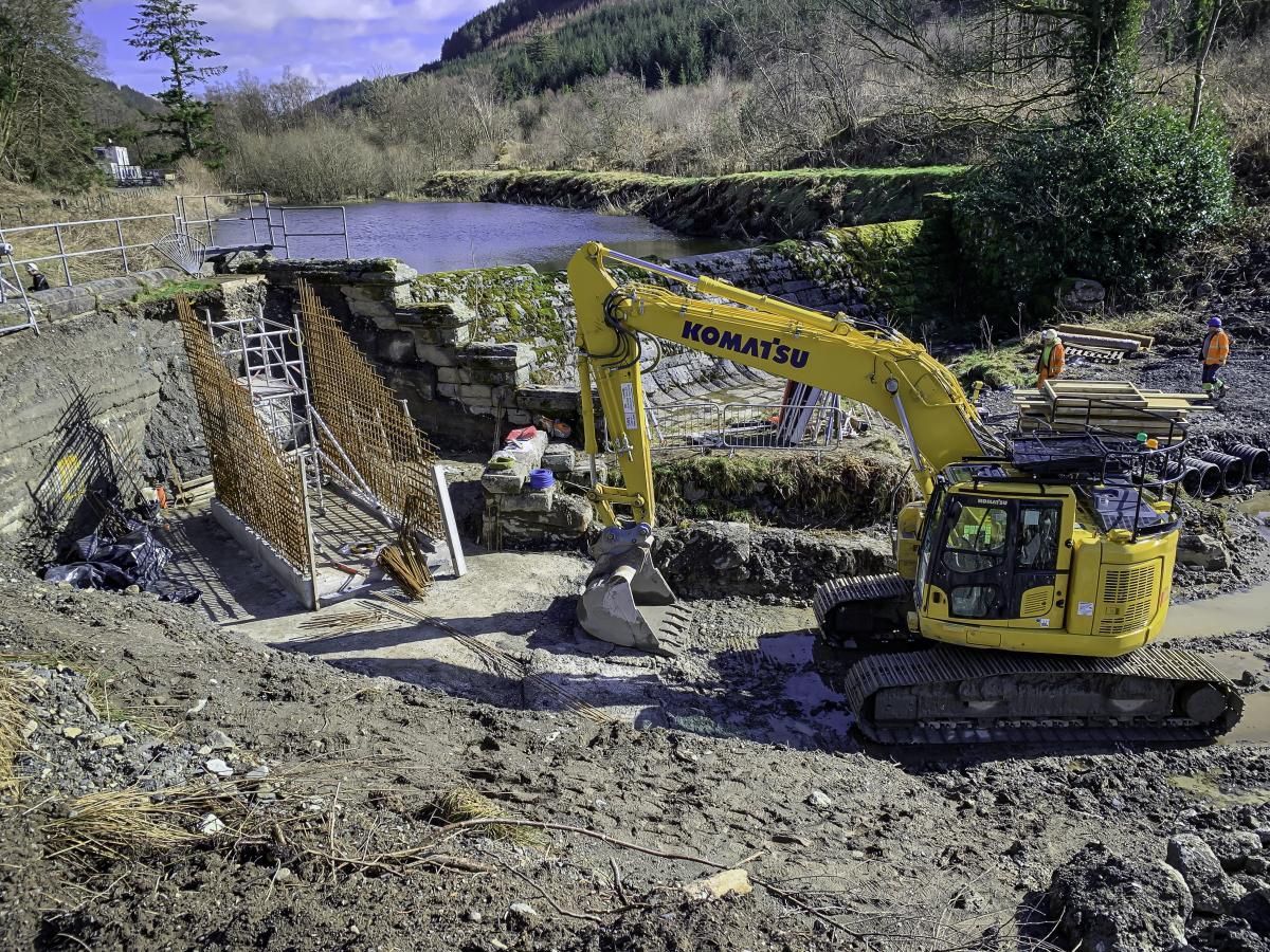 Yellow excavator at a construction site near a river. Rusty rebar and stone retaining walls are visible.
