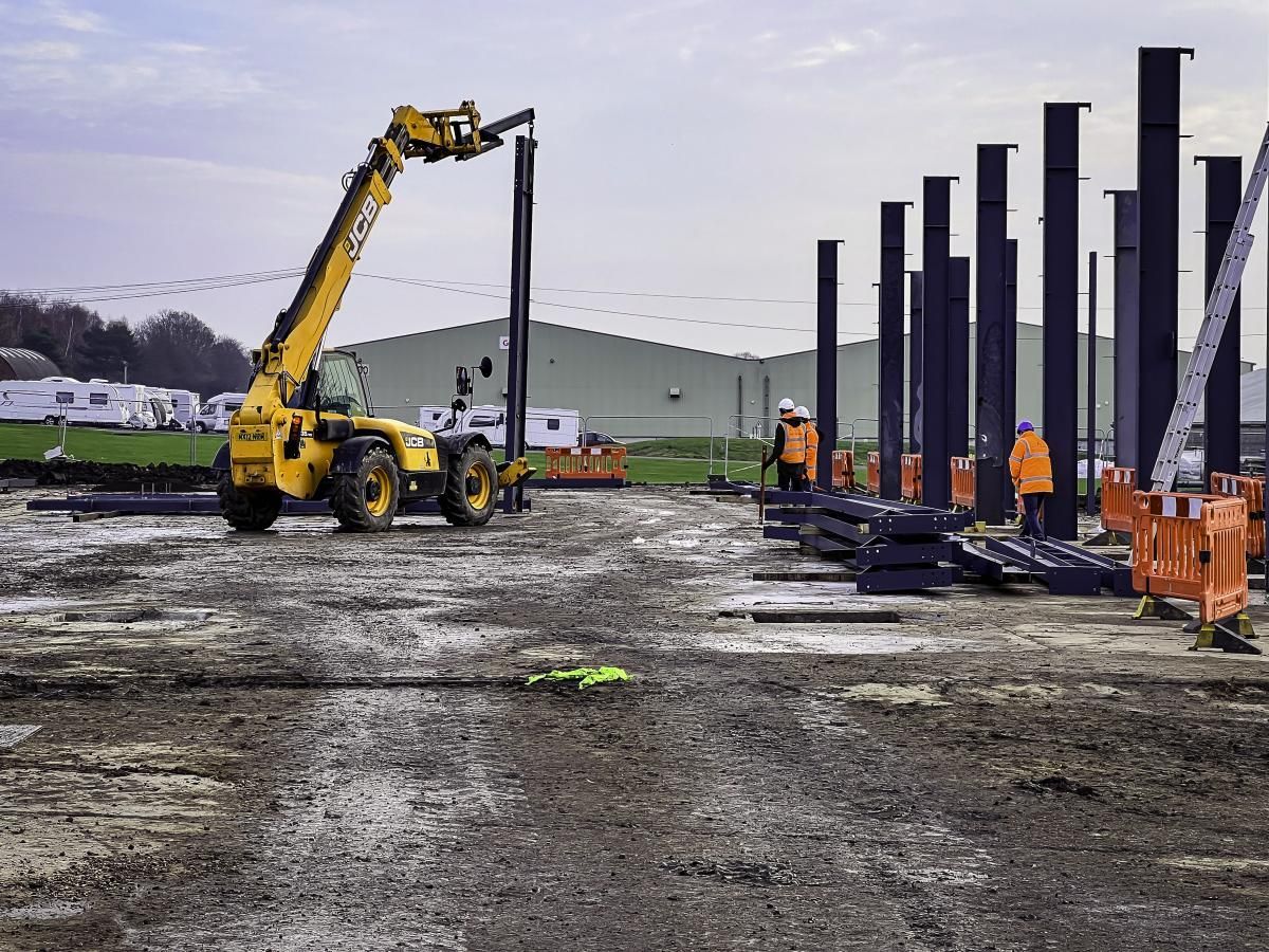Construction site with yellow telehandler lifting steel column. Workers in orange vests nearby.