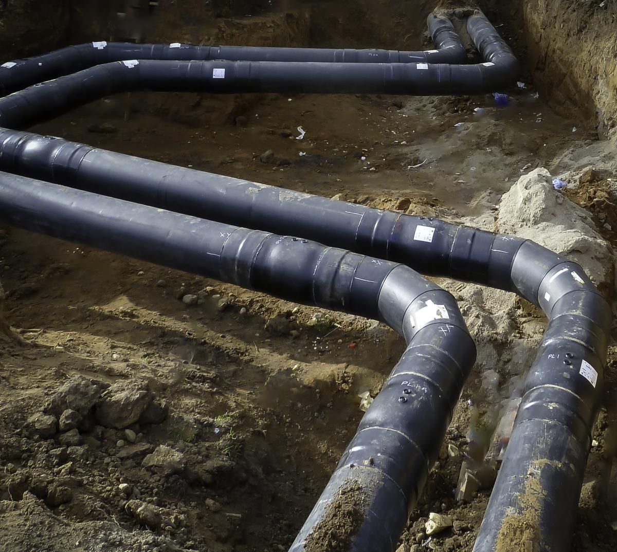 Black pipes in a trench, connected by curved joints, awaiting burial.