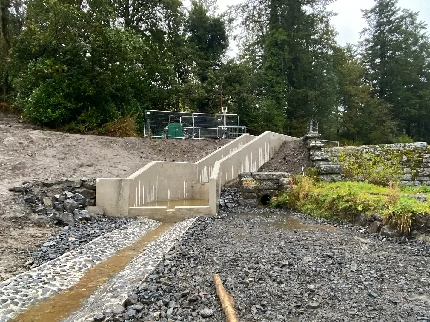 Concrete steps leading up a slope, with a drainage channel at the bottom. Gravel surrounds. Trees in the background.