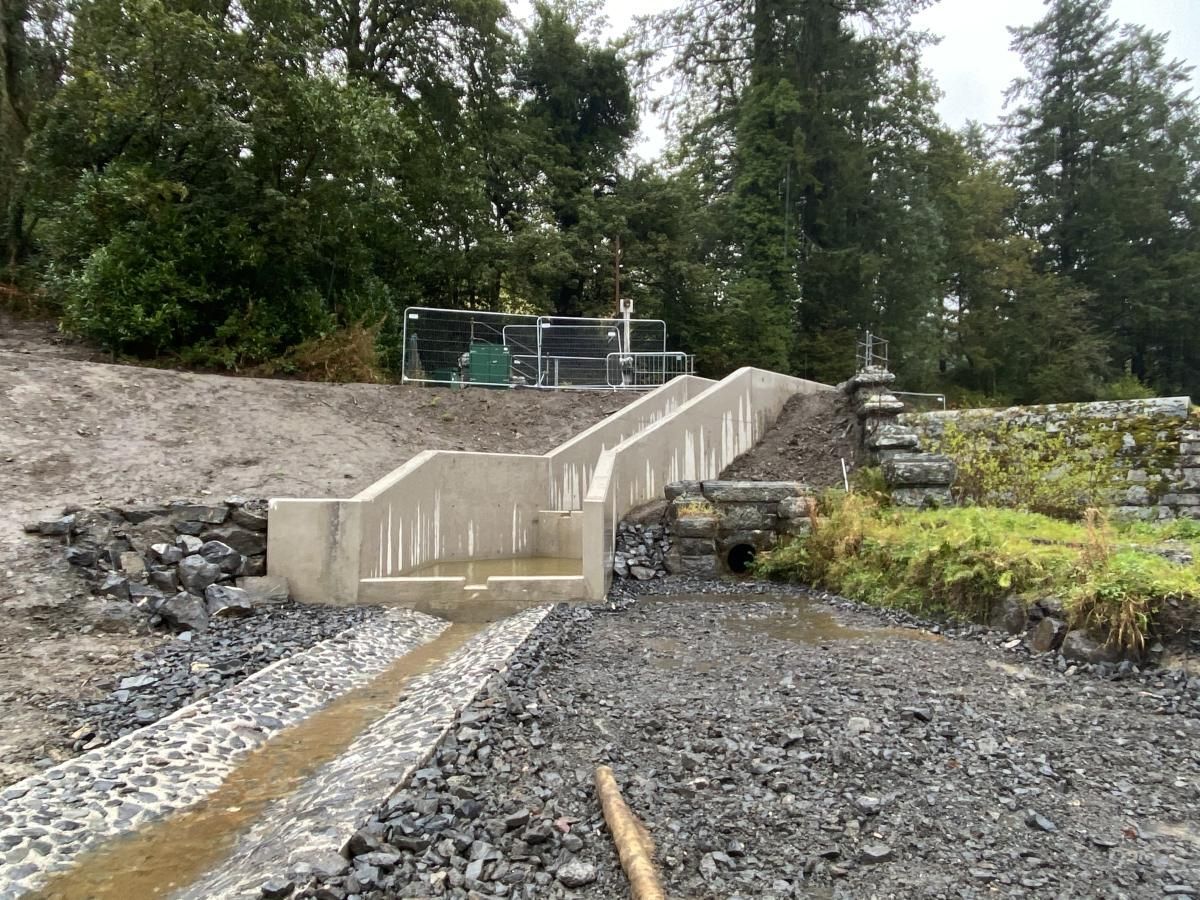 Concrete structure with water flowing downhill, gravel path, and trees in the background.