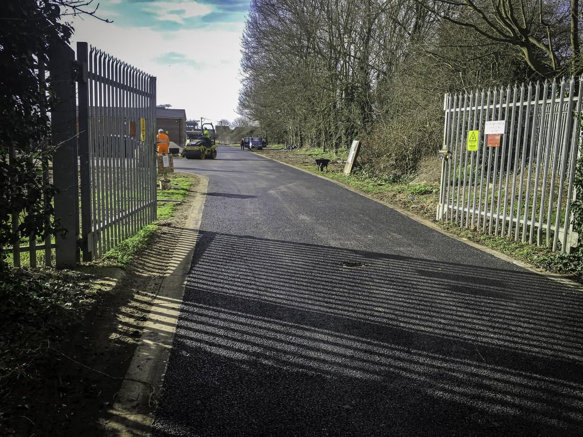 Black paved road leads into an industrial area, flanked by metal gates and greenery; sunny day.