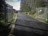 Black paved road leads into an industrial area, flanked by metal gates and greenery; sunny day.