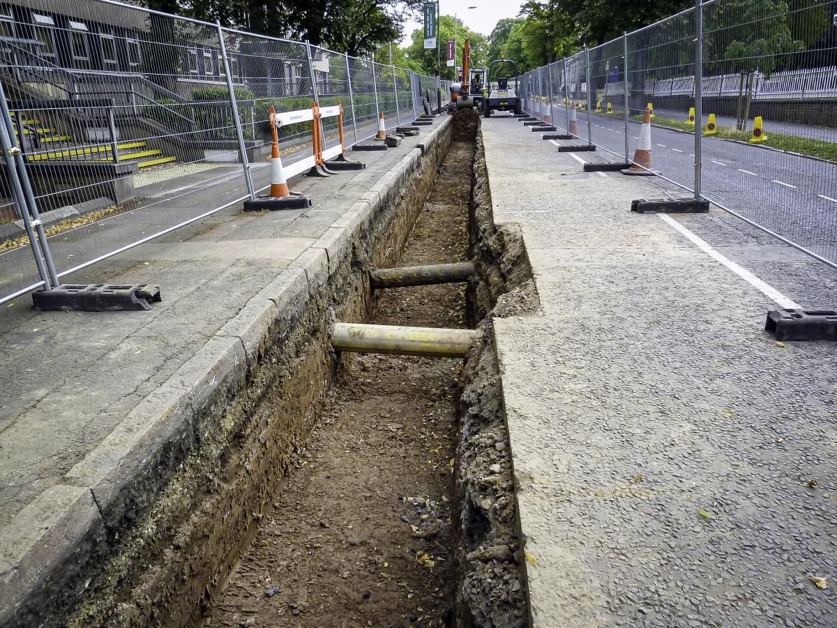 Trench dug in asphalt road, lined with fencing and cones. Wooden supports span the trench.