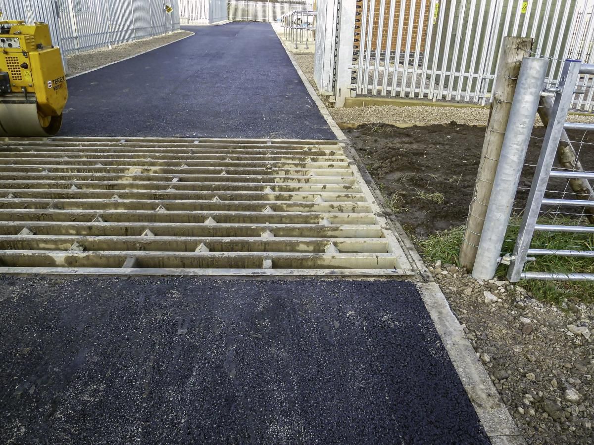 Asphalt paving over a grated drain, next to a fence and gate, with a small roller in the background.