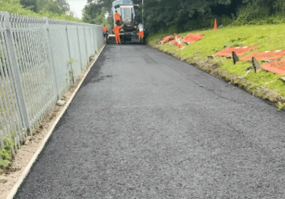 Paving crew laying asphalt on a road, beside a metal fence and grassy embankment.
