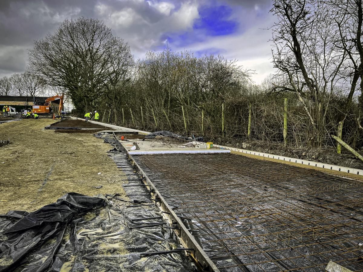 Construction site with rebar framework, dirt, and concrete forms, trees in the background under a cloudy sky.