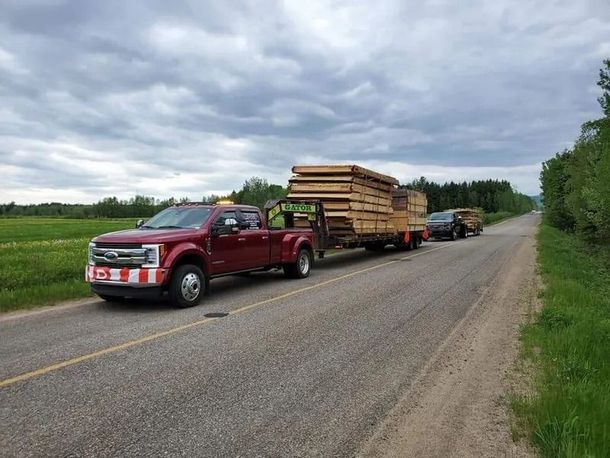 Un pick-up rouge tractant une remorque chargée de bois sur une route de campagne sous un ciel nuageux.