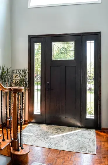 Dark wood front door with glass panels, patterned rug, and staircase, in a well-lit entryway.