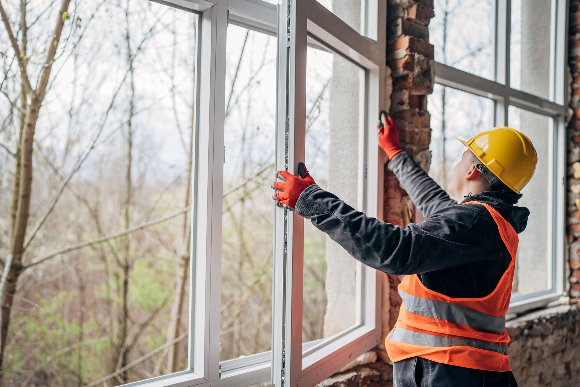 A worker in a hard hat and vest installs a window in a building with exposed brick.