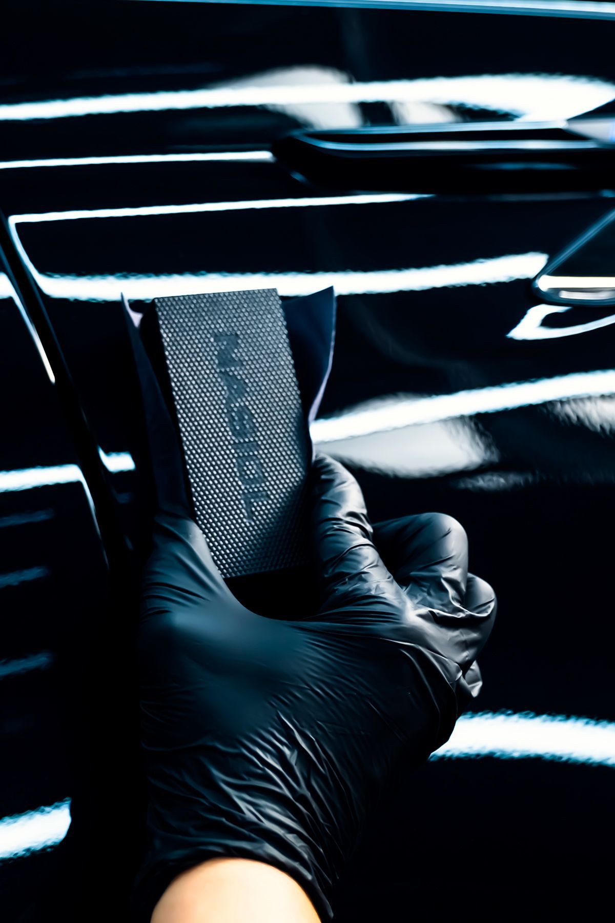 A person wearing black gloves is holding a sponge in front of a car.