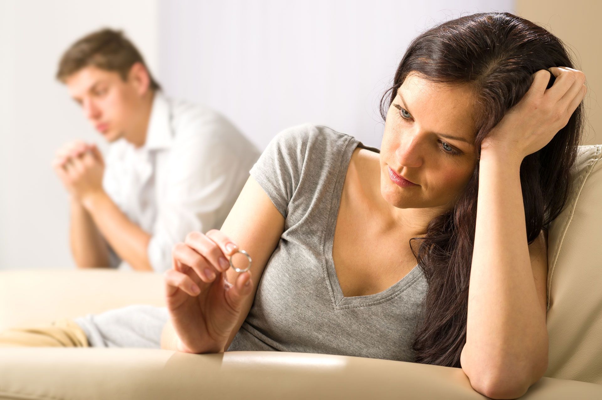 Woman lying on bed with head in hand, looking upset, while a blurred man sits in the background.