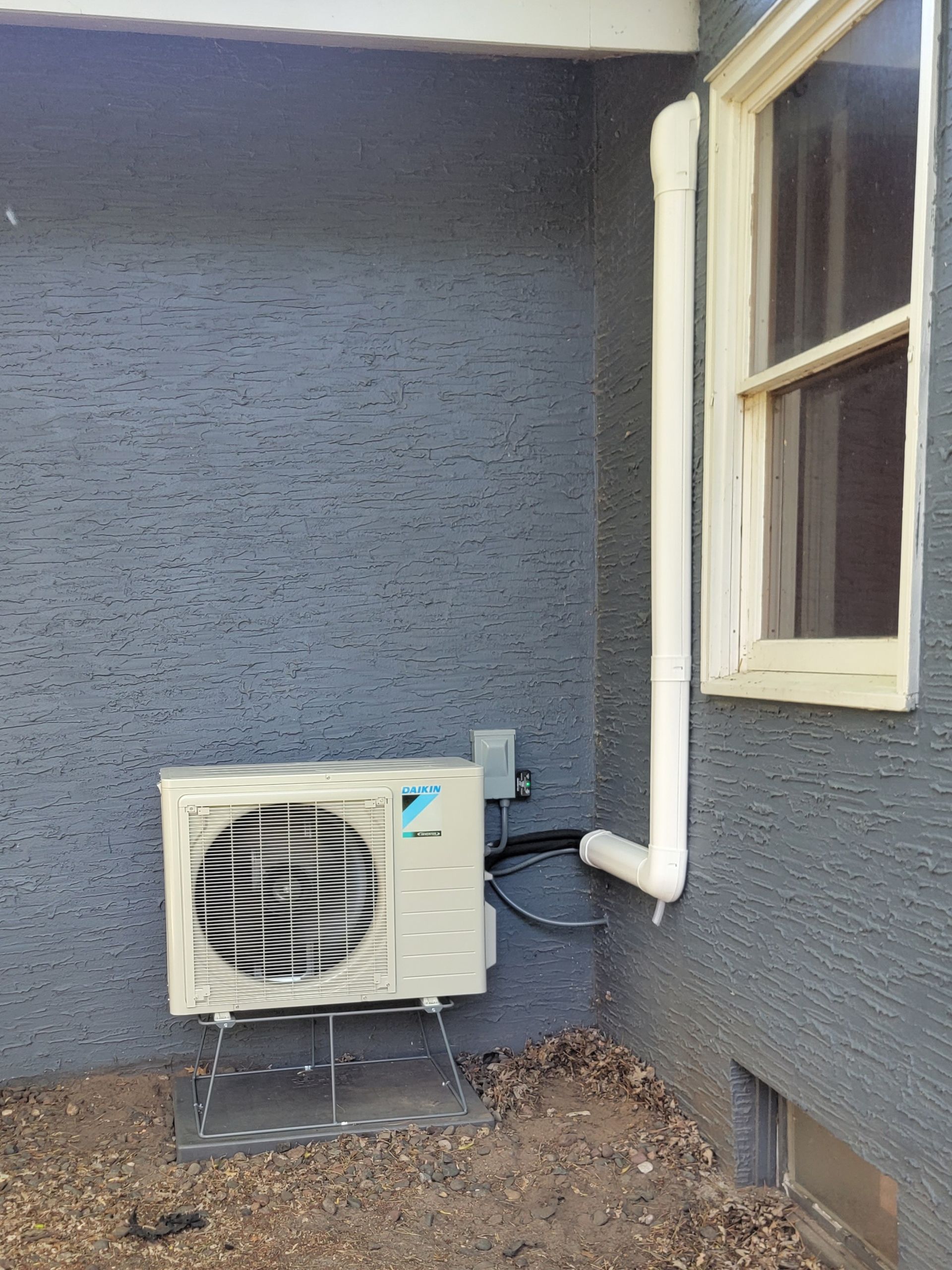 Outdoor air conditioning unit on a grey stucco wall next to a window and a drainpipe.