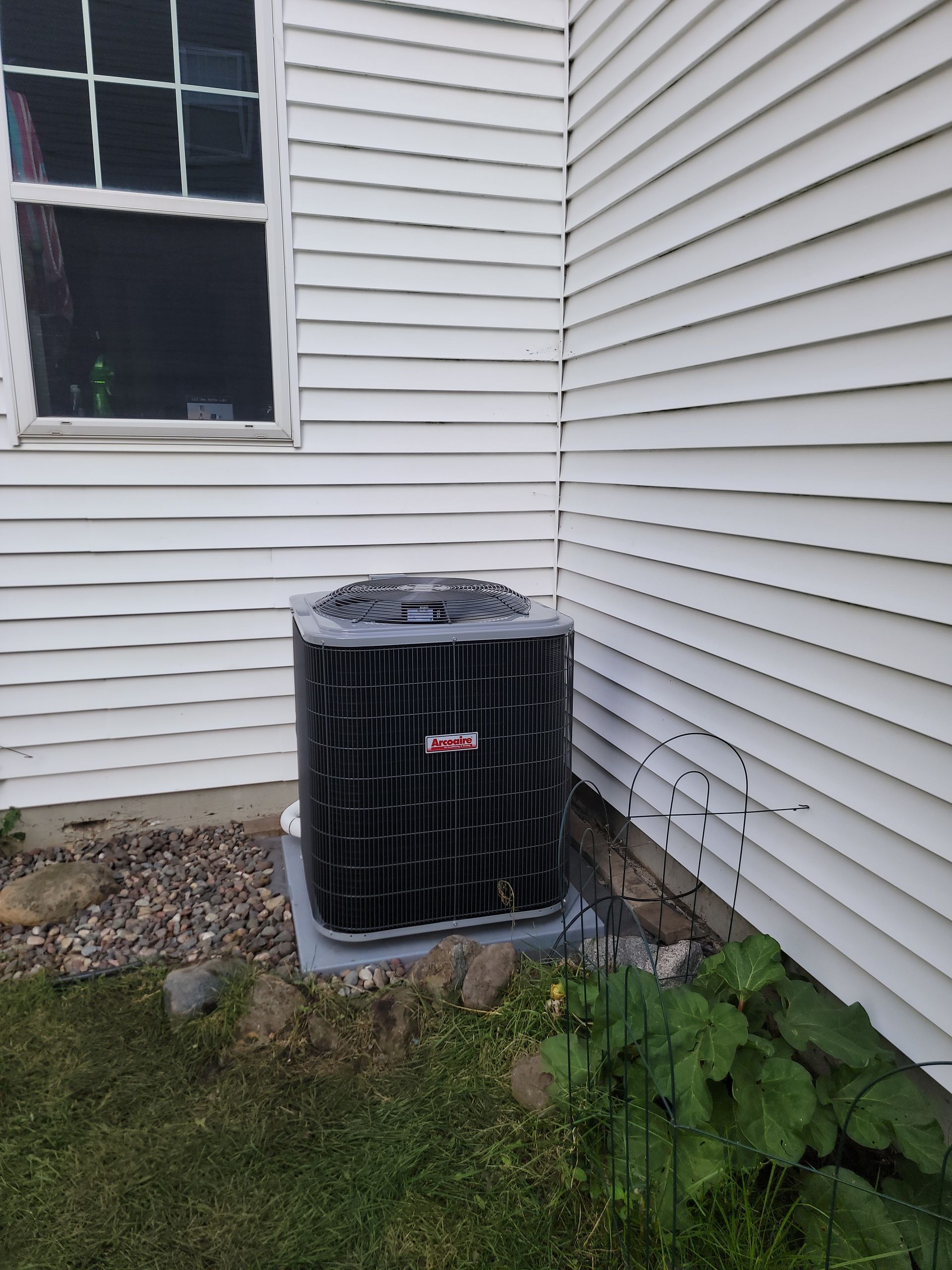 Air conditioner unit next to a white house with a window, set in grass and landscaping.