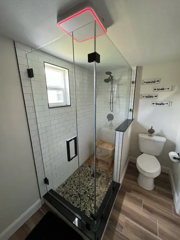 Modern bathroom with glass shower, white subway tile, and pebble floor. Overhead light with red glow, and wood-look flooring.