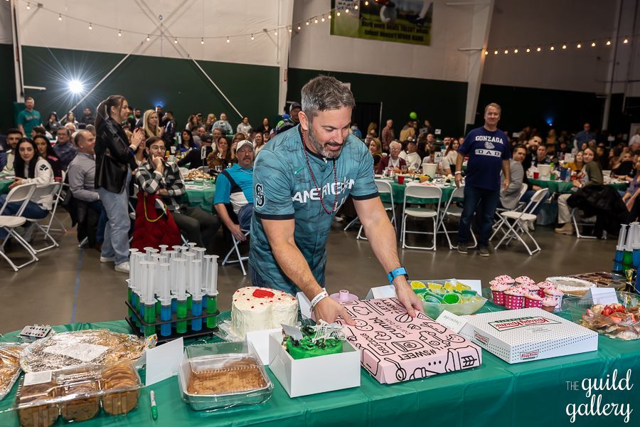 A man is cutting a cake at a party in front of a crowd.