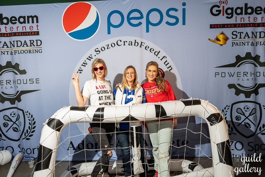 Three women are posing for a picture in front of a pepsi sign.