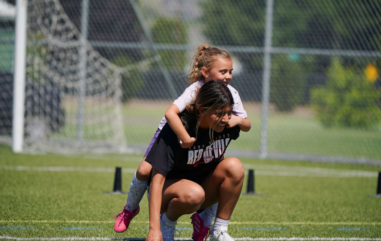 A woman is kneeling down with a little girl on her shoulders on a soccer field.