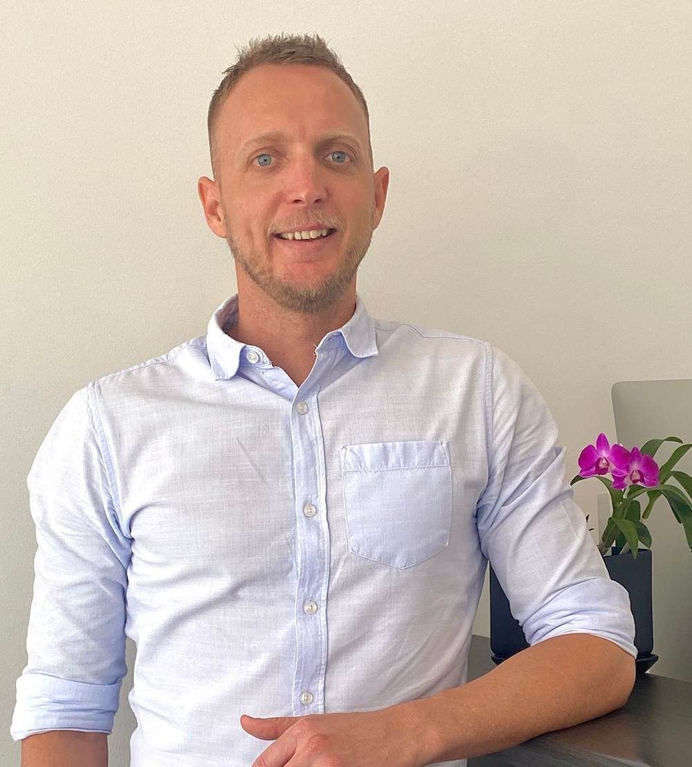 A Man In A White Shirt Is Sitting At A Desk With Flowers In The Background — Grays Chiropractic Centre In Rural View, QLD